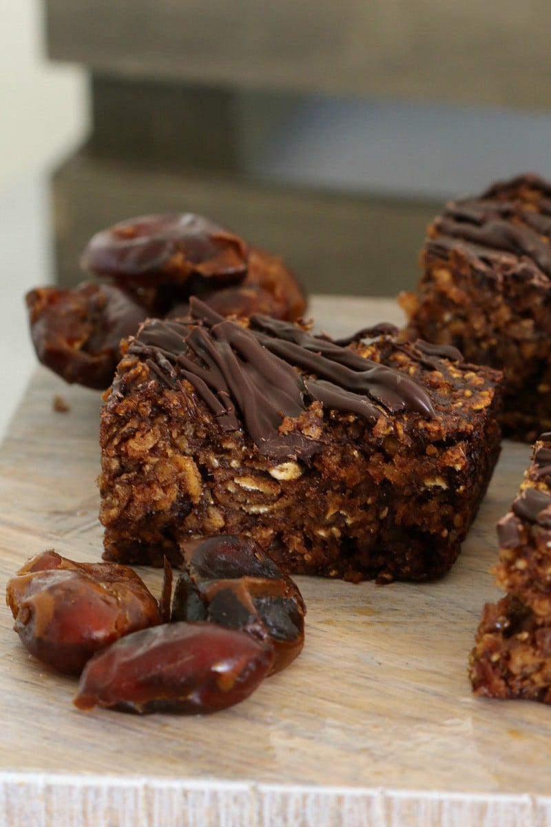 Pieces of a date and oat slice with dark chocolate on top, and some fresh dates on a wooden board