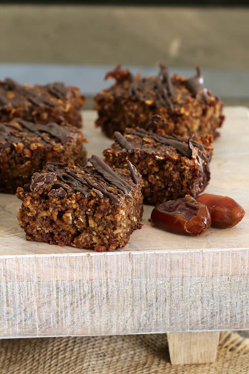 Squares of a date and oat slice drizzled with dark chocolate and two fresh dates beside them