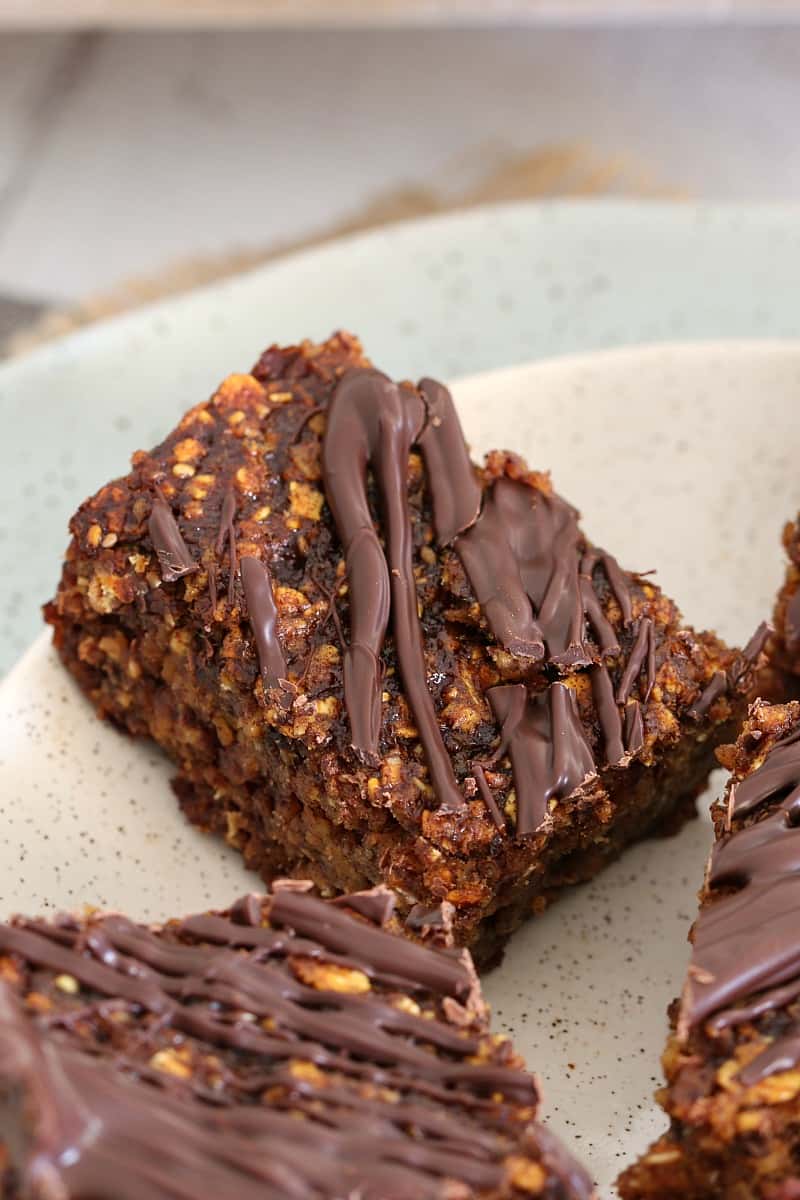 A close up pieces of a date and oat slice with dark chocolate drizzled on top served on a white plate