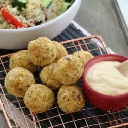 Healthy falafel balls piled on a copper wire tray with a bowl of hommus, in front of a bowl of quinoa salad