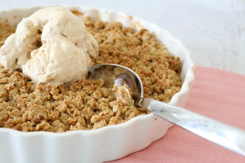 A dessert spoon in a white baking dish of apple crumble topped with ice cream
