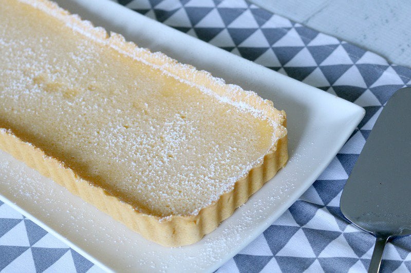 An oblong fluted lemon tart, dusted with icing sugar served on a white plate sitting on a blue and white place mat