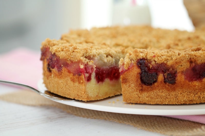 A cake with a layer of raspberries and a crumble topping on a white plate with one serve removed