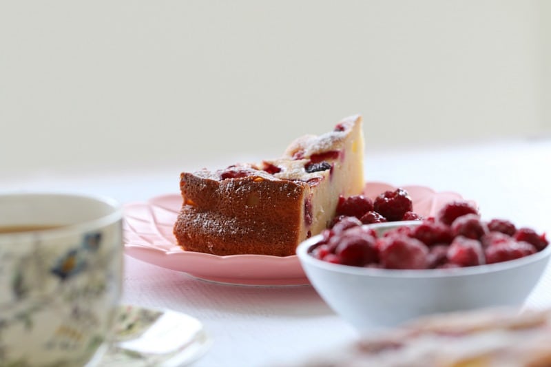 A serve of a raspberry ricotta cake on a pink plate next to a bowl of fresh raspberries