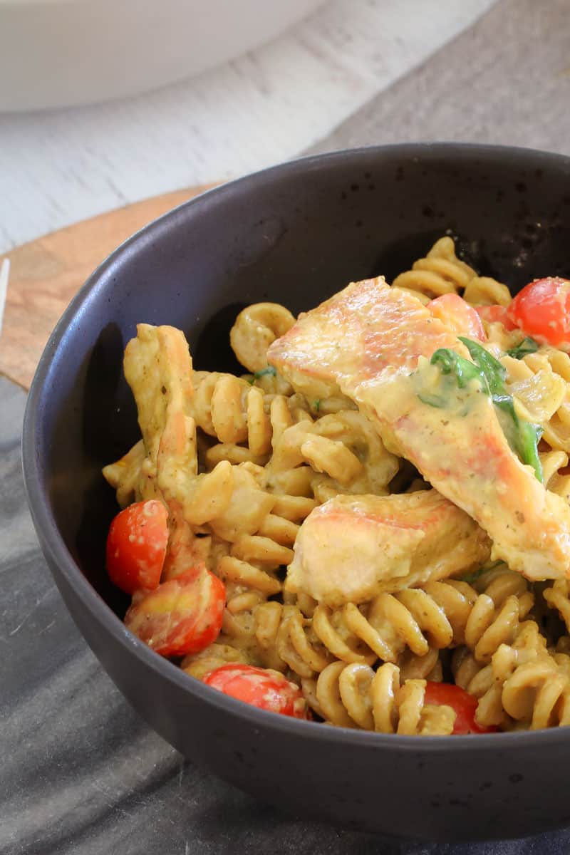 A close up of a black bowl filled with pasta, chicken, cherry tomatoes and spinach, in a creamy pesto sauce