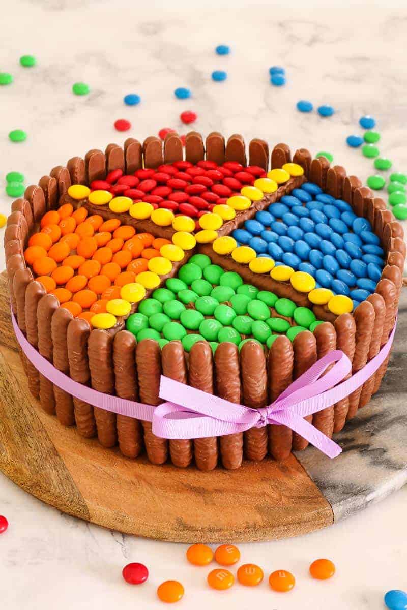 A round cake on a wooden board, decorated with chocolate log biscuits standing upright around the cake and coloured M&M\'s on top