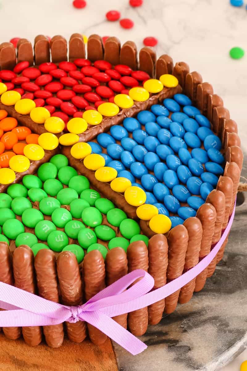 The side of a round cake with chocolate log biscuits around the outside, and coloured M&M\'s placed in the frosting on top