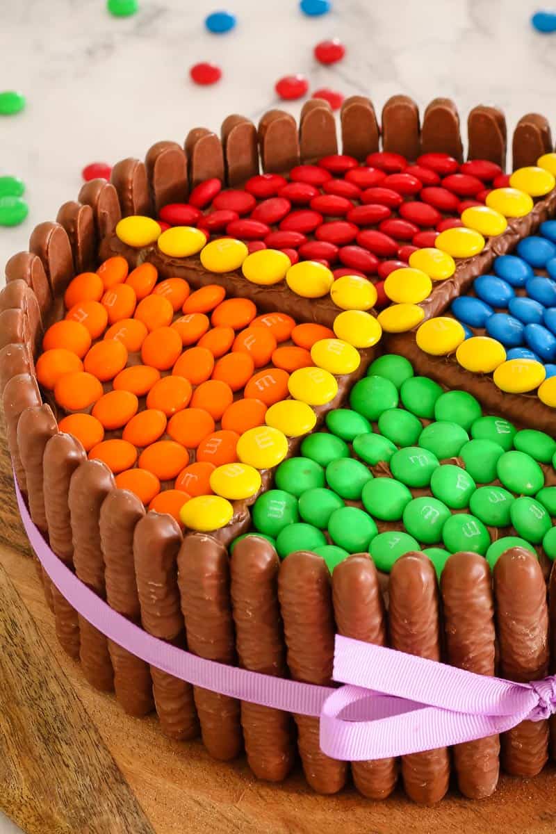 Coloured M&M\'s on top of a round cake that has chocolate log biscuits standing upright around the outside of the cake