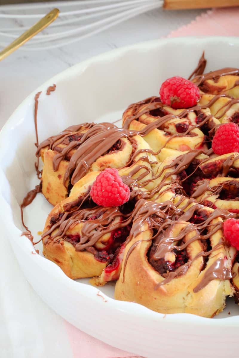 A white baking dish filled with scrolls drizzled with Nutella and topped with fresh raspberries