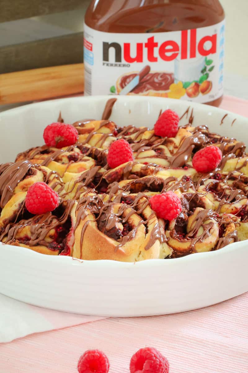 A round white baking dish filled with baked scrolls drizzled with Nutella and fresh raspberries on top