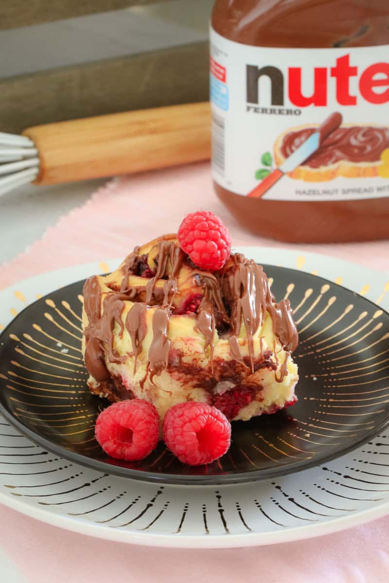 A scroll drizzled with Nutella served on a plate with fresh raspberries, in front of a jar of Nutella
