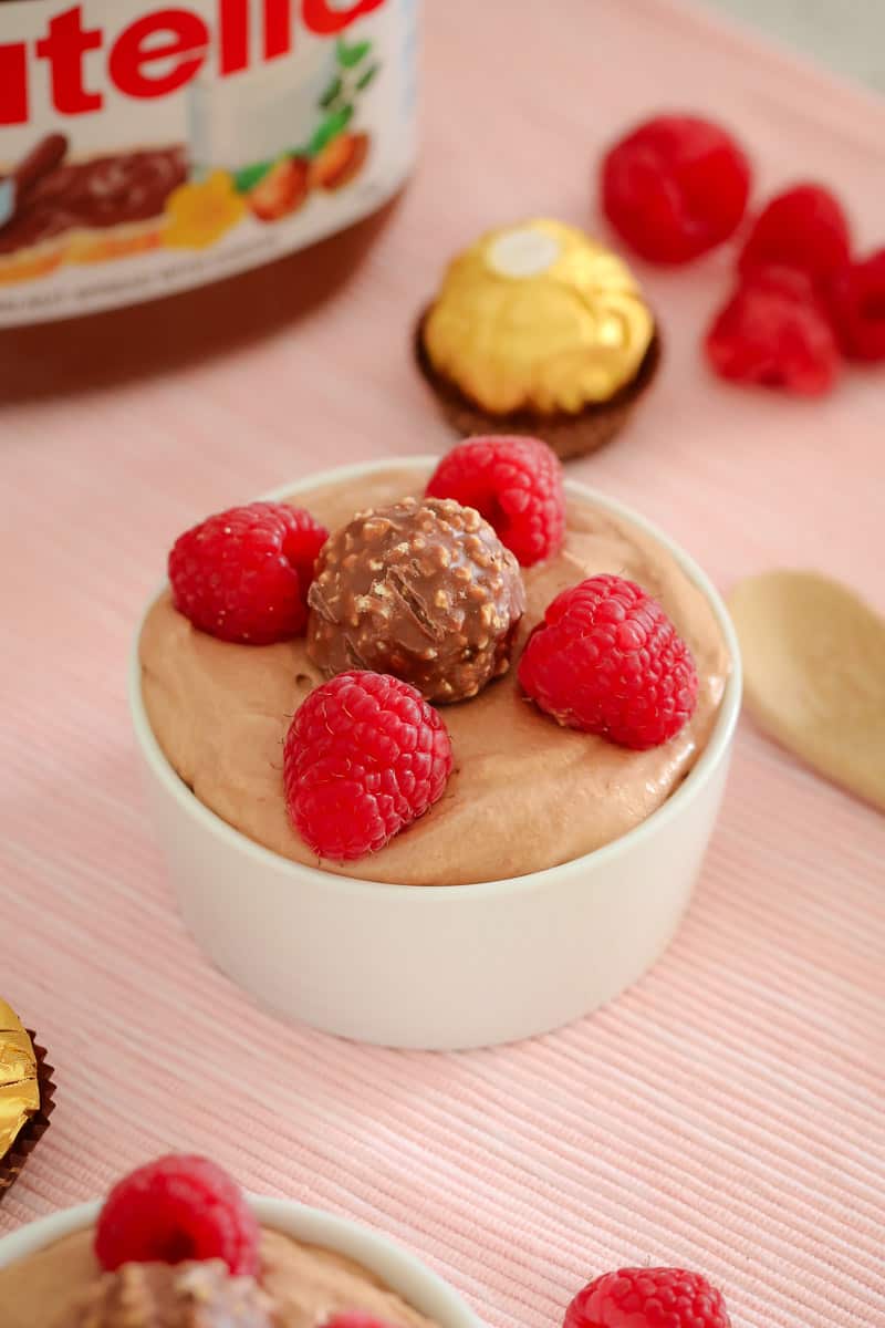 A small white bowl on a bench, filled with mousse and decorated with fresh raspberries and a Ferroro Rocher