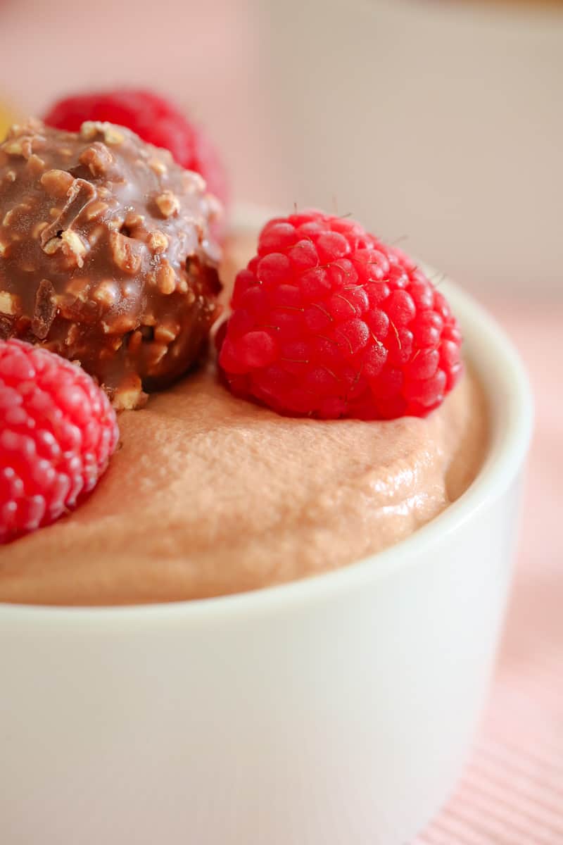 A close up of fresh raspberries and a Ferroro Rocher on top of mousse in a small white bowl