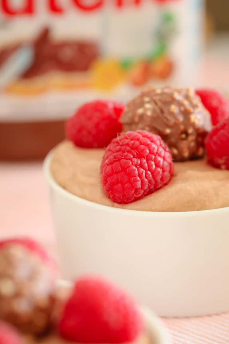 A close up side view of a small white bowl filled with mousse and decorated with fresh raspberries and a Ferroro Rocher