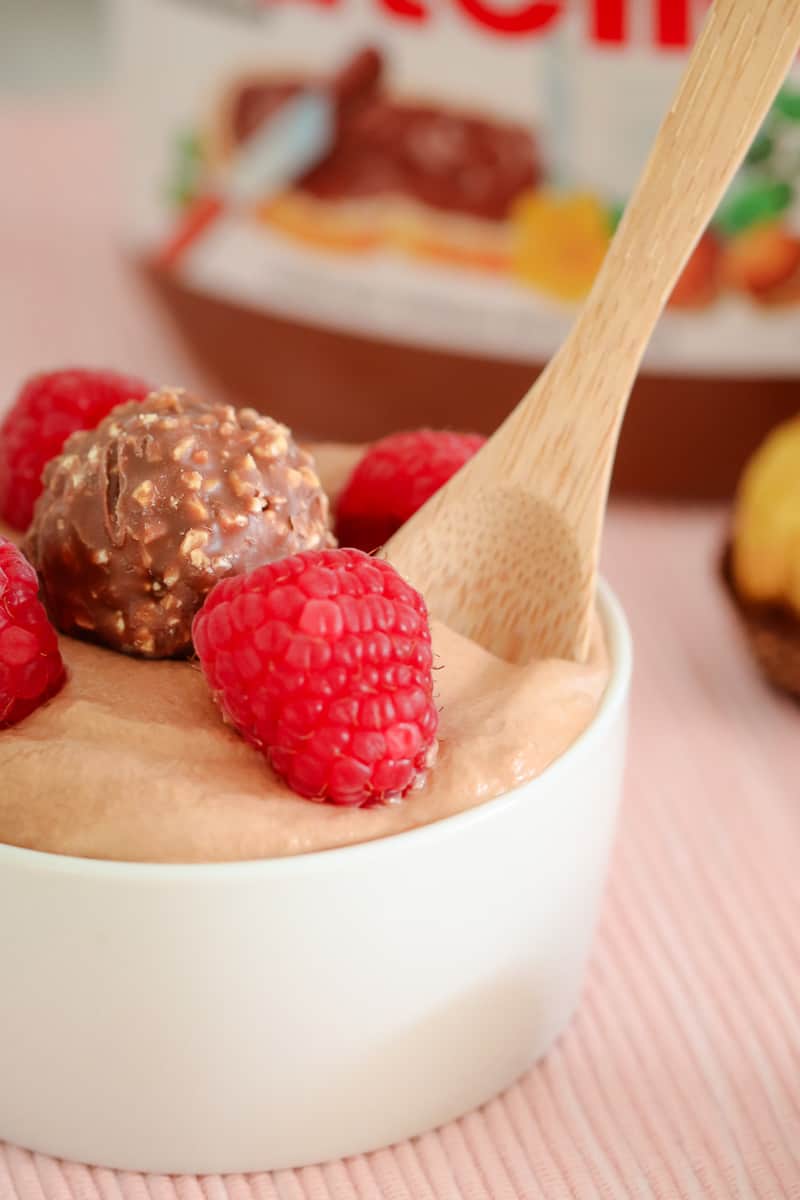 A wooden spoon in a small white bowl of mousse with fresh raspberries and a Ferroro Rocher