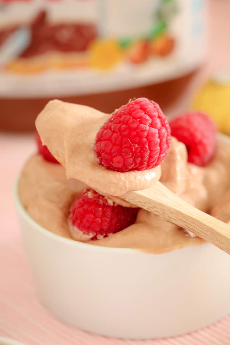 A wooden spoonful of mousse and a raspberry above a small white bowl filled with more mousse and raspberries