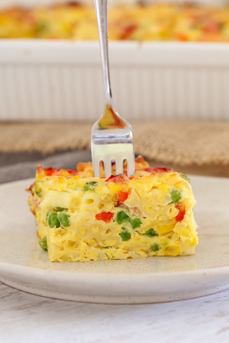 A fork standing up in a serve of frittata on a white plate, showing a filling of peas, corn and capsicum