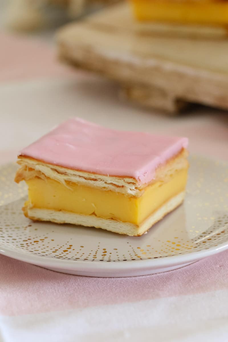 A square slice made with a pastry top and base, a pink icing glaze and filled with vanilla custard.