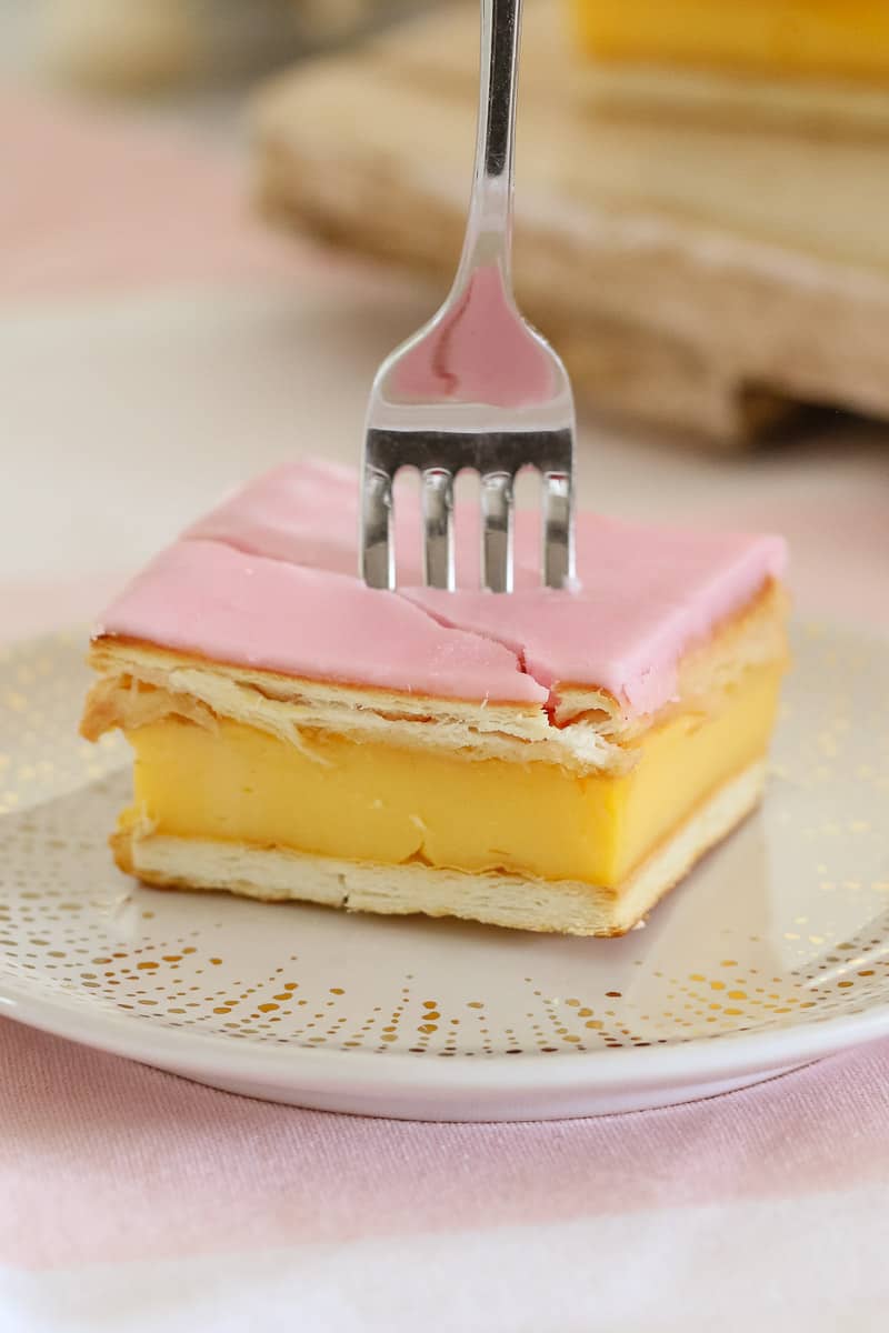 A fork pressed into a square slice of custard and pastry with  pink icing on a plate.