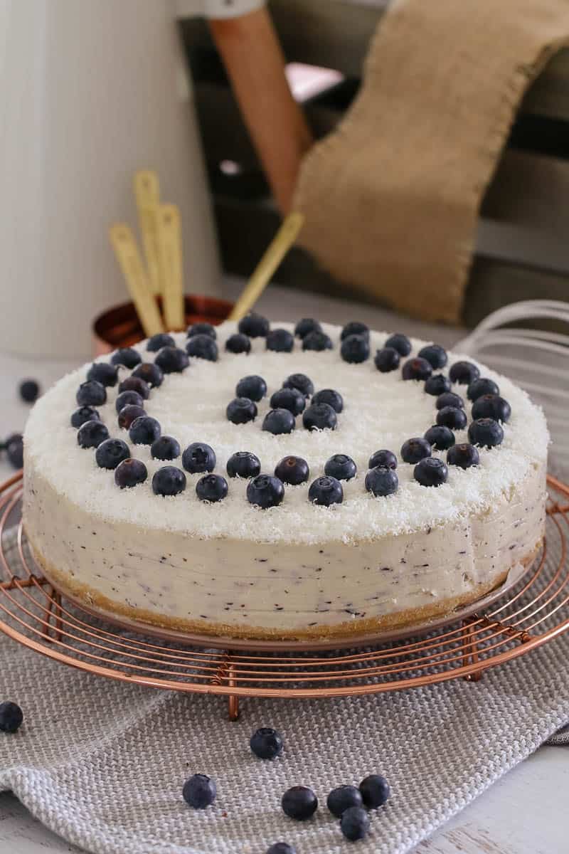 A white cheesecake topped with fresh blueberries on a copper wire tray