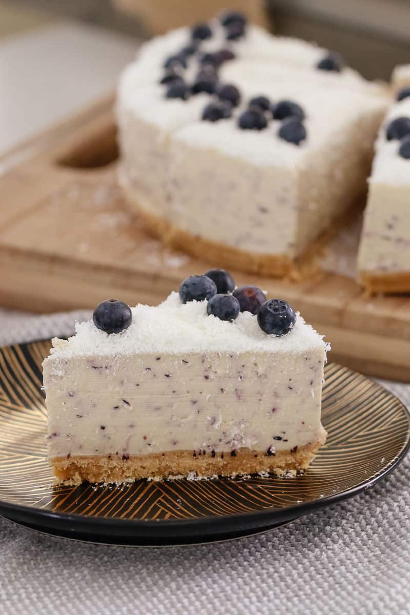 A serve of cheesecake topped with fresh blueberries on a plate in front of the remaining cheesecake