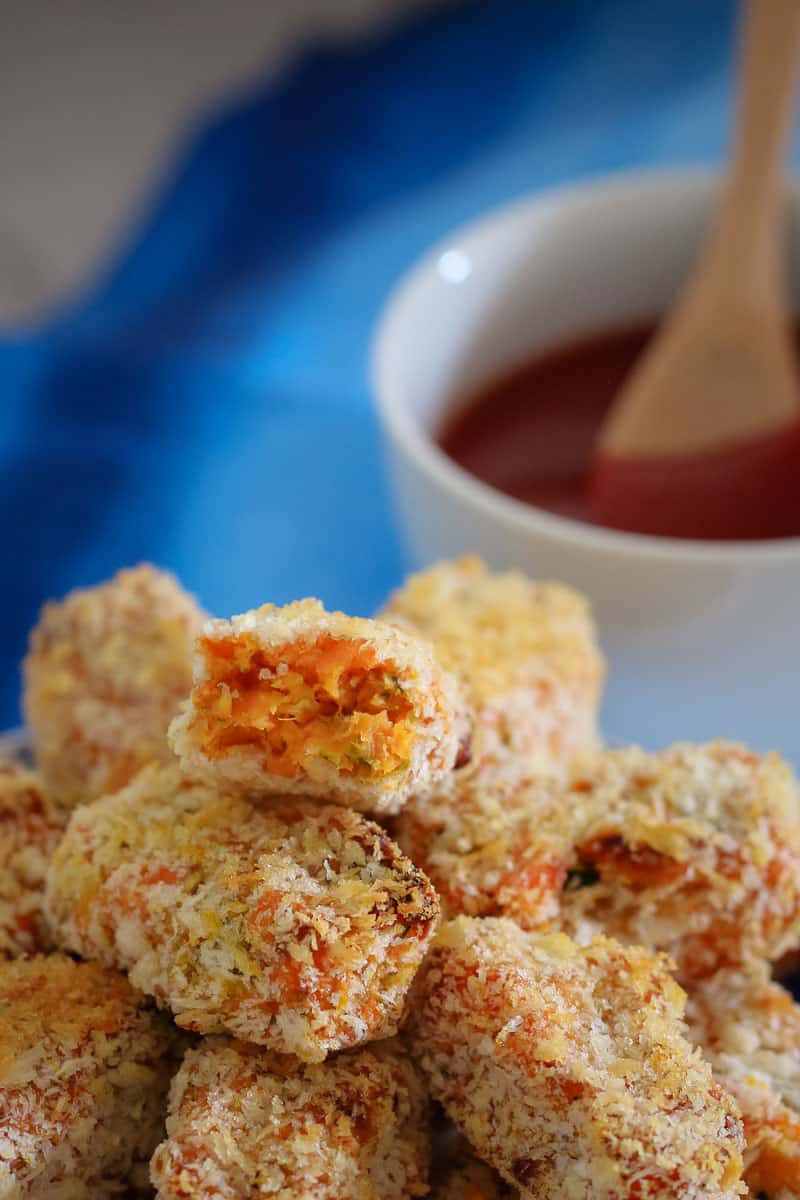 A close up of bread crumbed nuggets with one split to show filling, and a bowl of sauce behind