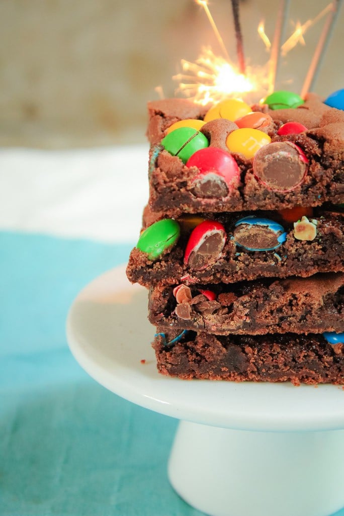 Pieces of a chocolate slice topped with coloured M&M's and stacked on a white cake stand
