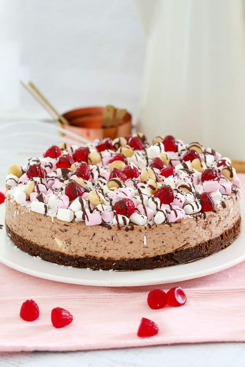 A chocolate cheesecake topped with marshmallows, raspberries, nuts and drizzled with chocolate on a white plate