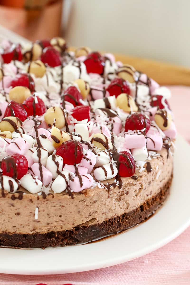 A close up of a rocky road chocolate cheesecake topped with marshmallows, raspberries, nuts and drizzled with chocolate
