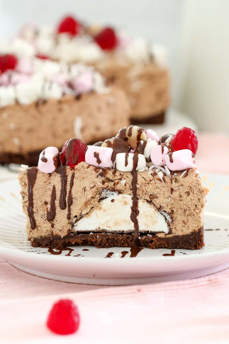 A side view of a serve of rocky road cheesecake showing filling and topped with marshmallows, raspberries, nuts and drizzles of chocolate