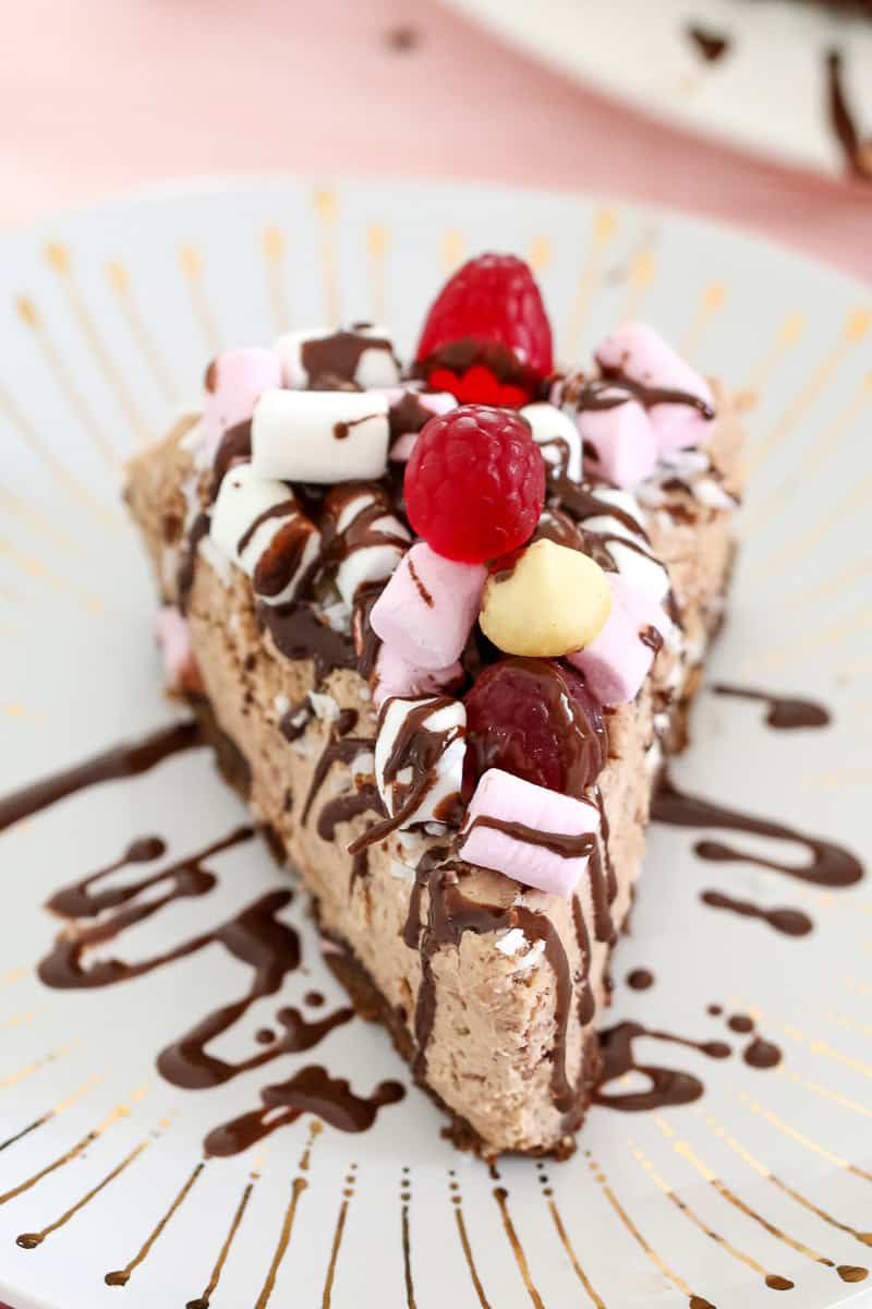 A serve of chocolate cheesecake on a plate, topped with marshmallows and raspberries, and drizzled with chocolate