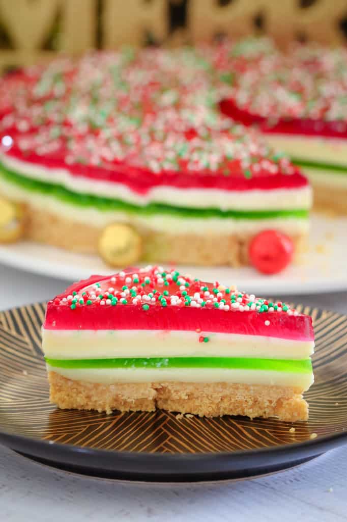 A close up serve of red, white and green layered Jelly Cake, topped with sprinkles, on a plate, with remainder of cake in background