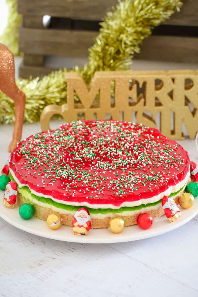 A round Jelly Cake made with green, white and red layers, and sprinkles on top, surrounded with Christmas decorations and chocolates