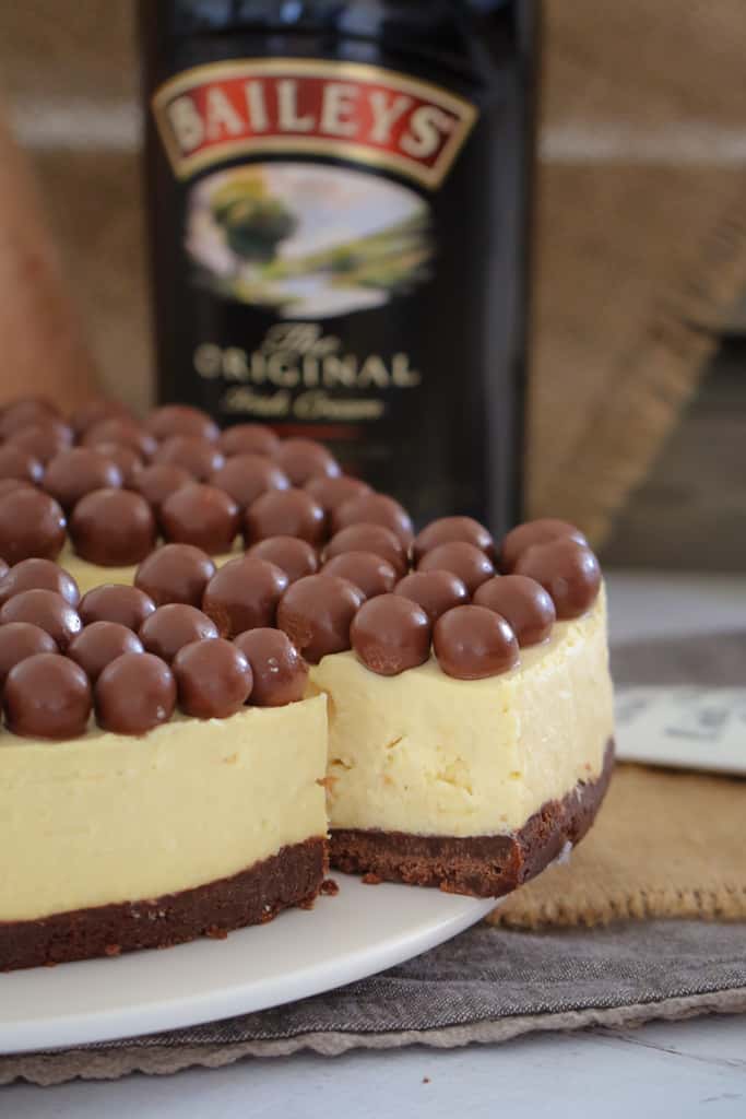 A white cheesecake with a chocolate base, and topped with Malteser balls with one slice cut and partially removed