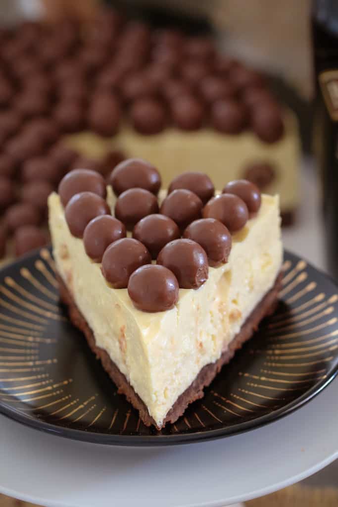 A serve of white cheesecake with a chocolate base, topped with Malteser balls on a black and gold plate