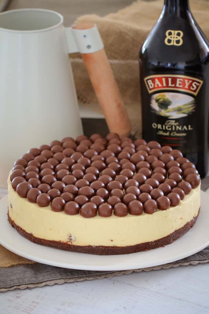 A white round cheesecake topped with Malteser balls on a white serving plate in front of a bottle of Baileys