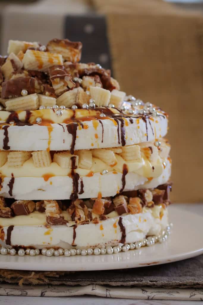 A close up of a three layered pavlova with layered of filling, drizzled with chocolate and caramel, and topped with chopped chocolate bars and wafer biscuits