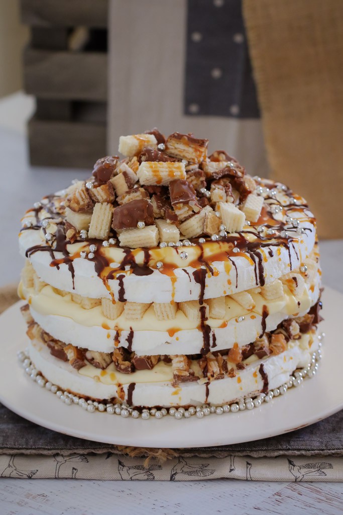 A three layered pavlova, with cream filling, drizzled with chocolate and caramel, and topped with chopped Picnic bars and wafer biscuits, on a serving plate