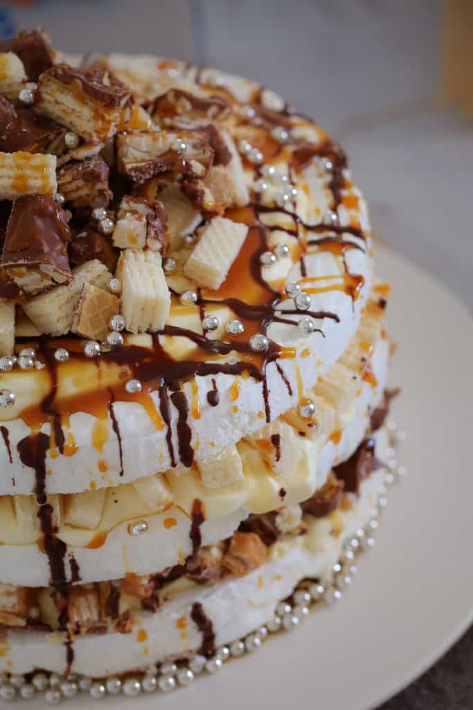 A close up of a three layered pavlova with fillings, drizzled with chocolate and caramel and topped with chopped Picnic bars and wafer biscuits