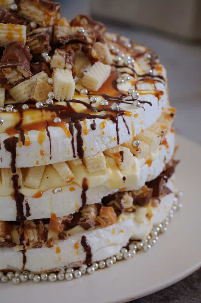 A close up side view of a three layered pavlova with layers of filling, drizzled with chocolate and caramel, and topped with chopped PIcnic bars and wafer biscuits