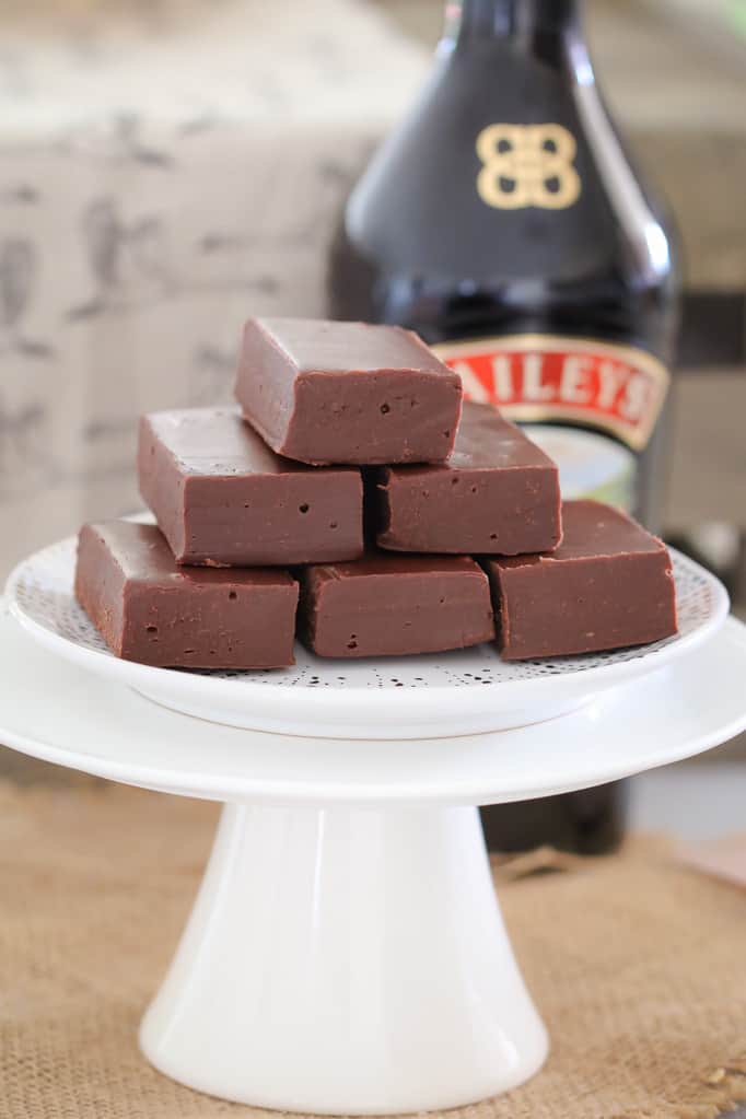A stack of chocolate fudge pieces on a white cake stand with a bottle of Baileys behind