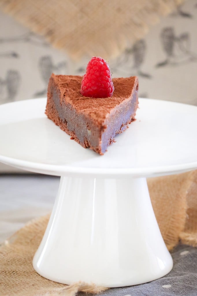 A slice of chocolate torte decorated with a fresh raspberry on a white cake stand