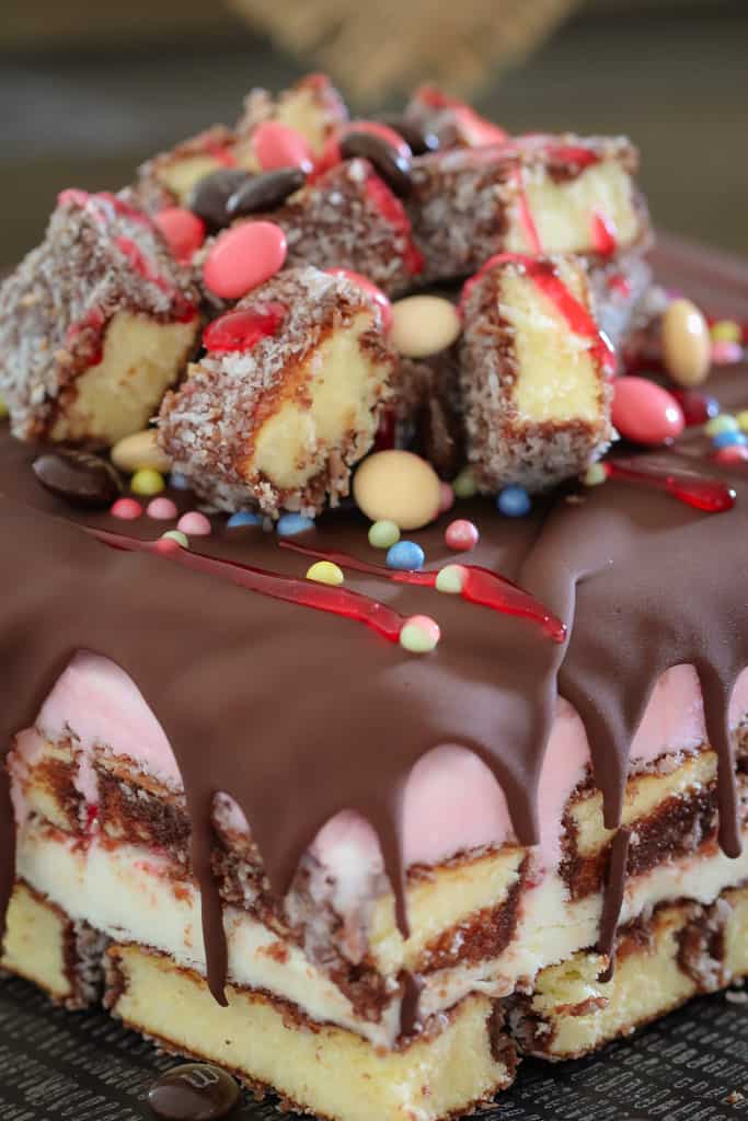 A close up of a layered lamington ice cream cake drizzled with chocolate and raspberry topping, and decorated with lamingtons and sweets