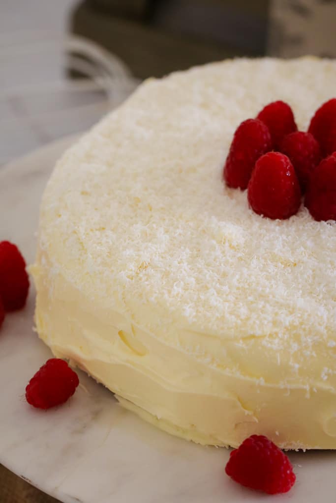 A white chocolate mud cake covered in white buttercream with a scattering of coconut and fresh raspberries to decorate