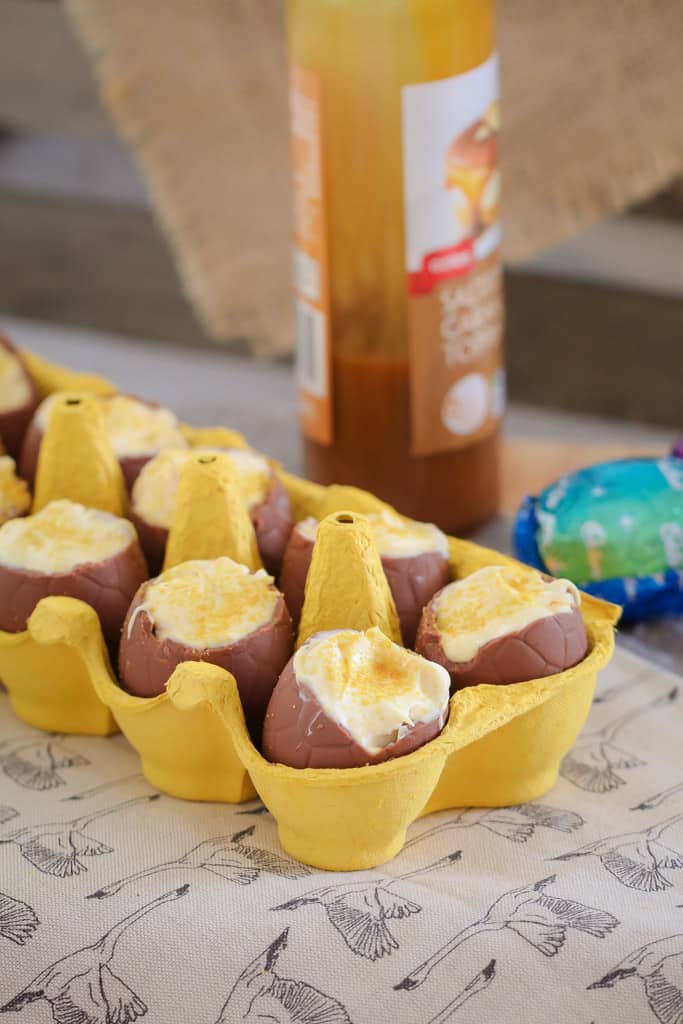 An egg carton filled with easter eggs and a bottle of caramel sauce in the background.