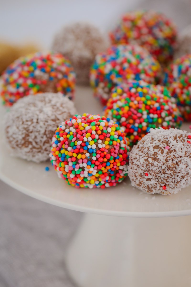 A white cake stand with round balls, some rolled in coconut and some in coloured sprinkles