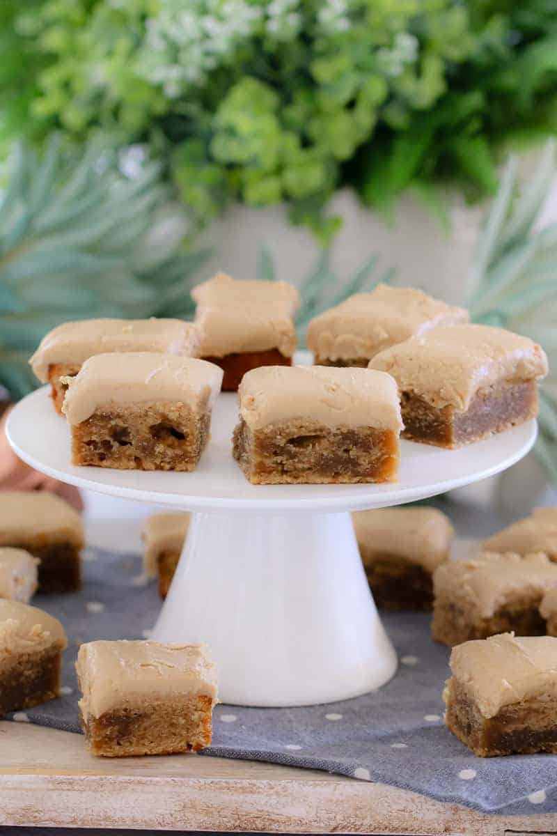 A bunch of squares of slice topped with caramel frosting with some on a white cake stand
