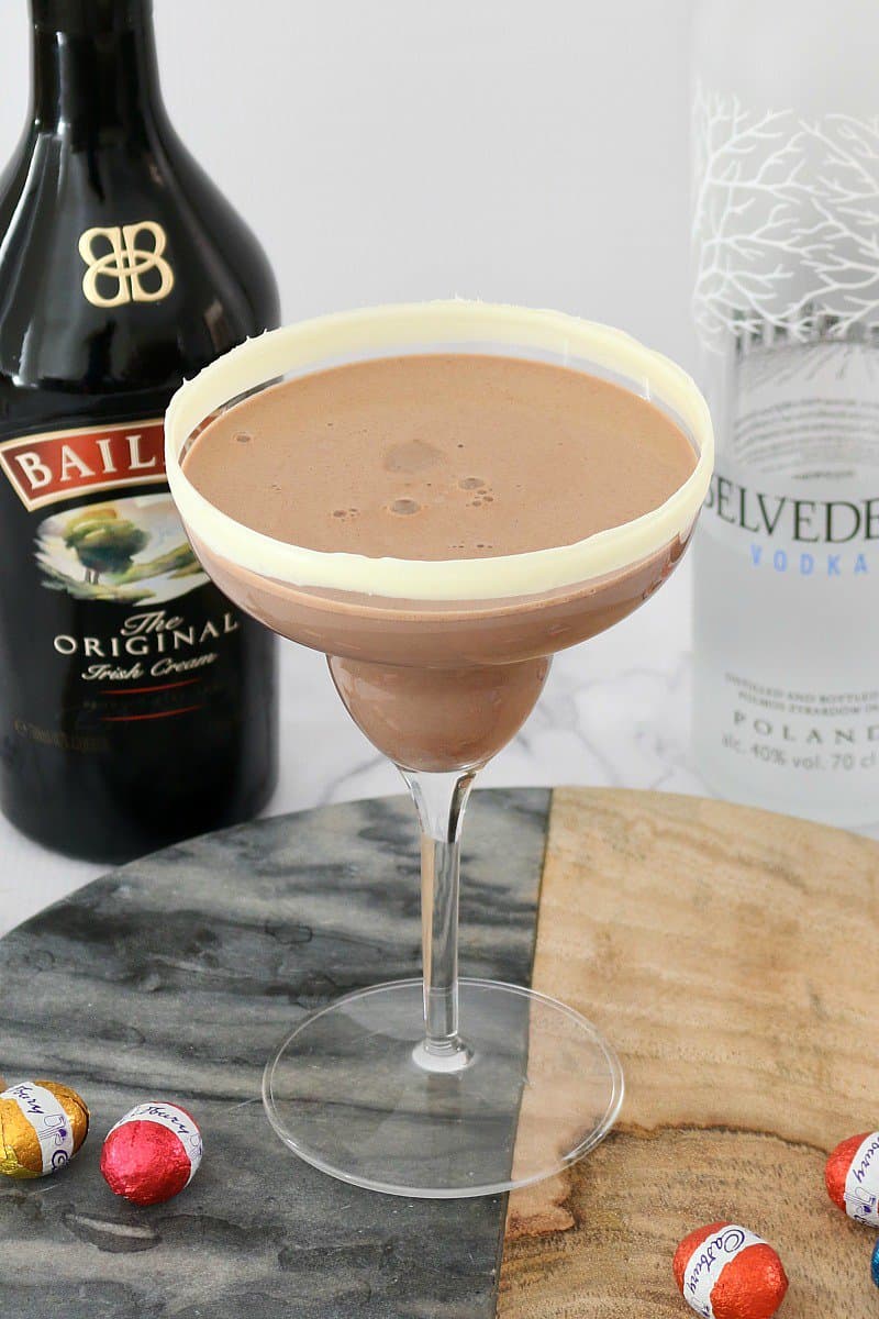 A long stemmed cocktail glass on a marble board, filled with a Baileys flavoured cocktail with a bottle of Baileys behind