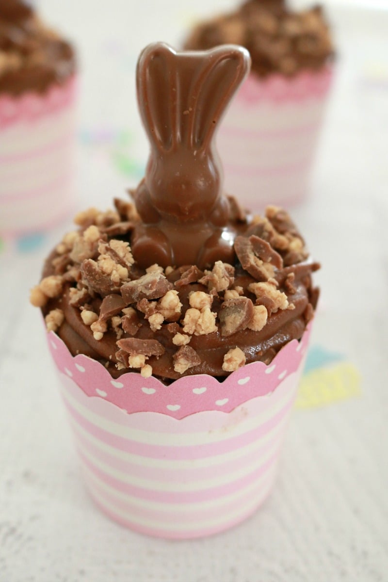 A chocolate cupcake in a pink paper cupcake holder decorated with chocolate frosting, crumbled Malteser\'s and a chocolate bunny on top