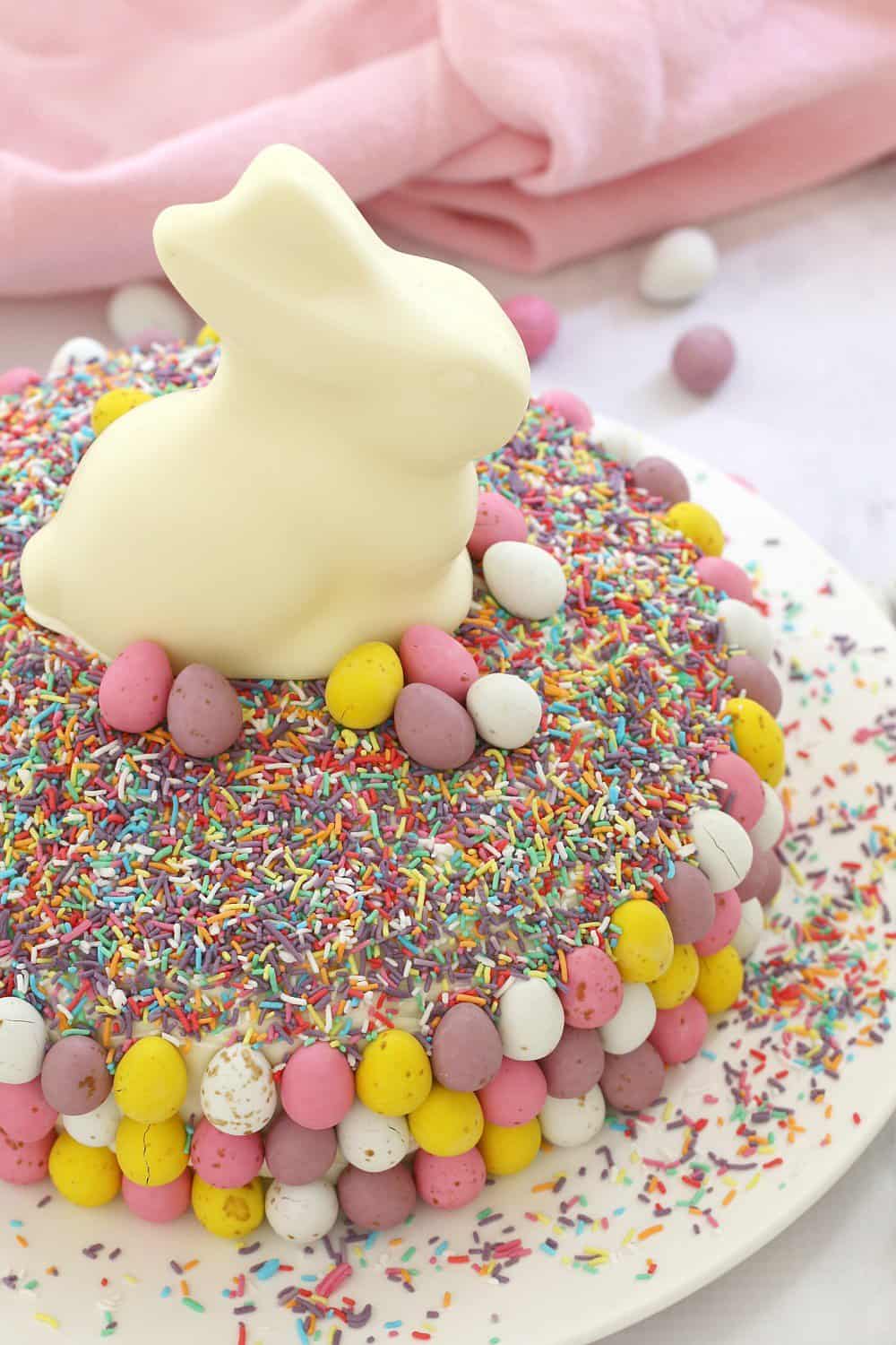 A round cake decorated with sprinkles, mini coloured Easter eggs and a white bunny on top